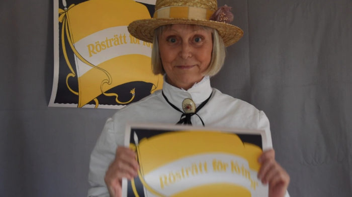 Eva Bergengren med hatt och blus enligt 1910-talets mode och två gula affischer som det står ”rösträtt för kvinnor” på.