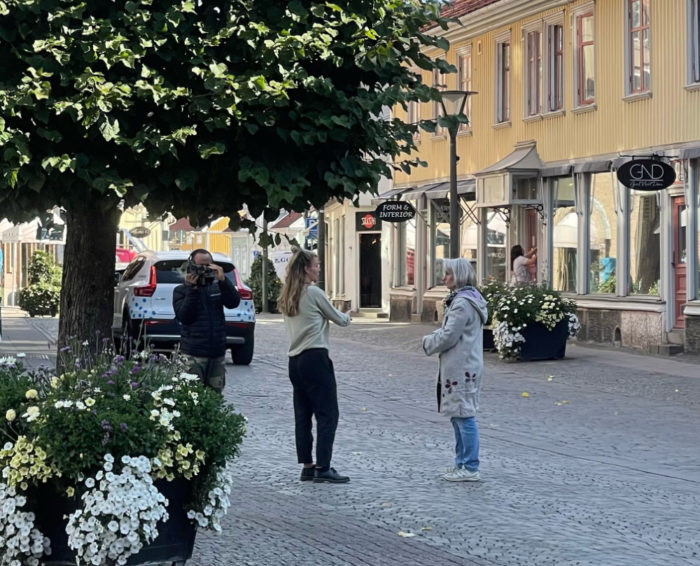 Eva Bergengren blir intervjuad av reporter och kameraman på Rådhustorget i Falkenberg. De står under ett träd med gul träbyggnad i bakgrunden.